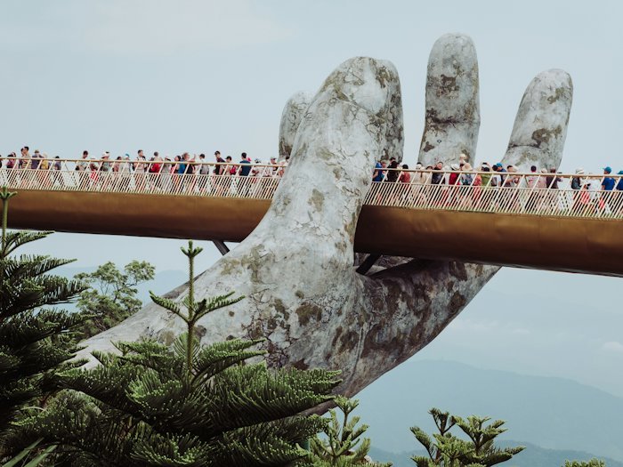 A footbridge held up by giant stone hands above forested hills